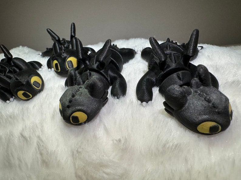 May include: Four black plush dragon toys with yellow eyes, lying on a white fluffy surface.