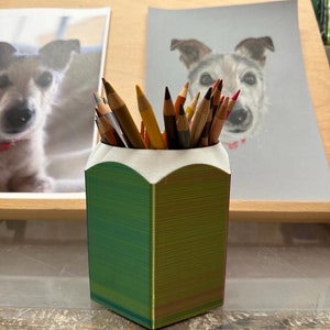 May include: A green and yellow pencil holder with a white rim, filled with coloured pencils. The holder is on a wooden surface with two drawings of dogs in the background.