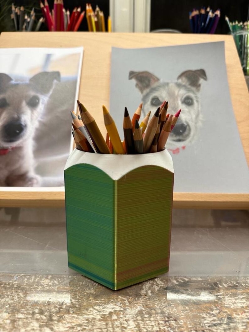 May include: A green and yellow pencil holder with a white rim, filled with coloured pencils. The holder is on a wooden surface with two drawings of dogs in the background.
