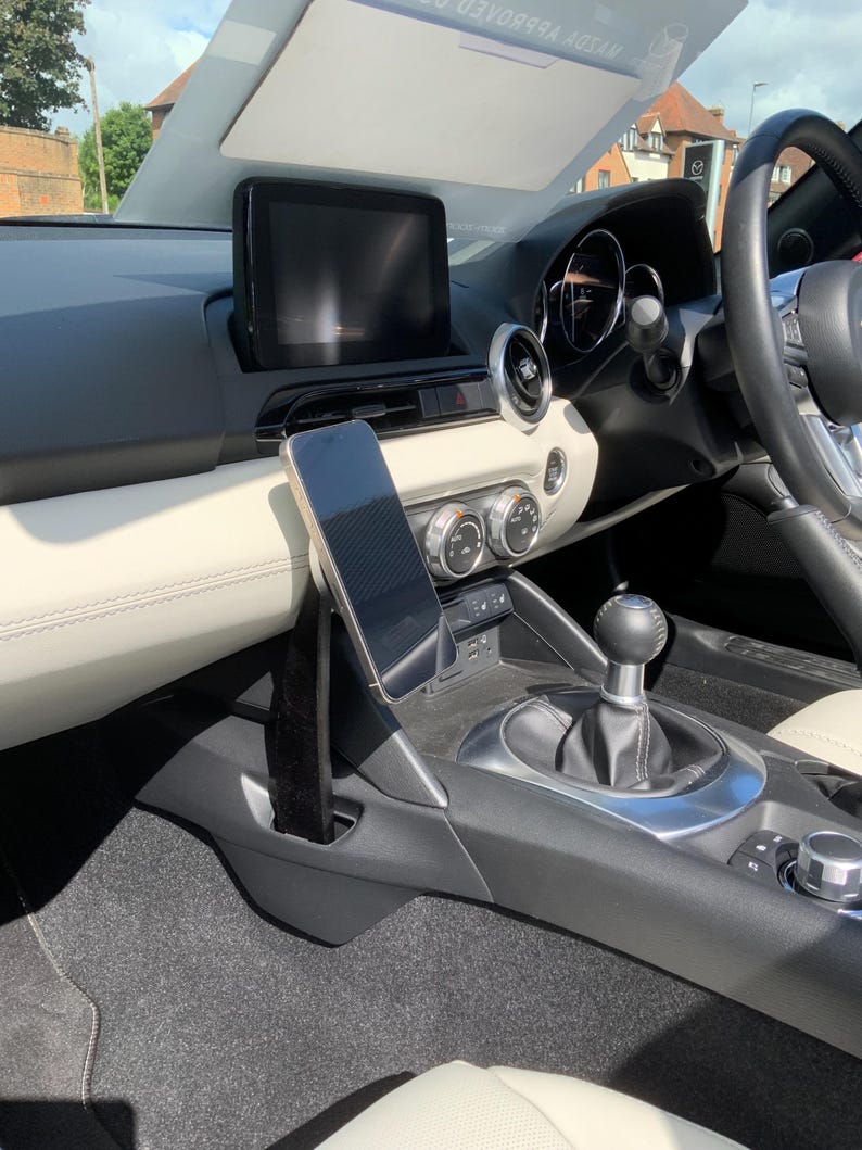 K&ouml;nnte beinhalten: Innenansicht eines Autos mit Smartphone-Halterung, zentralem Display und Bedienelementen. Das Armaturenbrett ist schwarz und cremefarben, mit Fokus auf Lenkrad und Schalthebel. Der Innenraum ist gut beleuchtet.