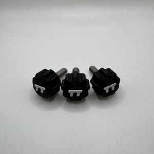 May include: Three black knobs with silver threaded shafts. Each knob has a textured surface and the letters "TT" are embossed on the top. The knobs are arranged in a horizontal line against a white background.