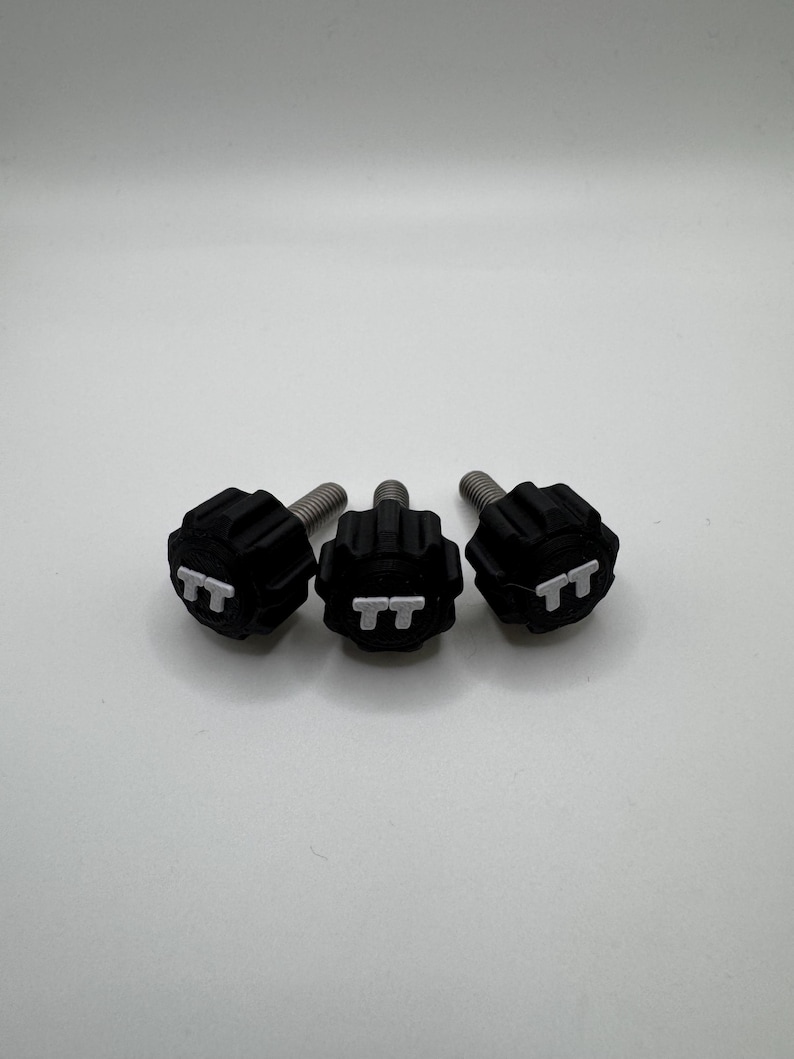 May include: Three black knobs with silver threaded shafts. Each knob has a textured surface and the letters "TT" are embossed on the top. The knobs are arranged in a horizontal line against a white background.