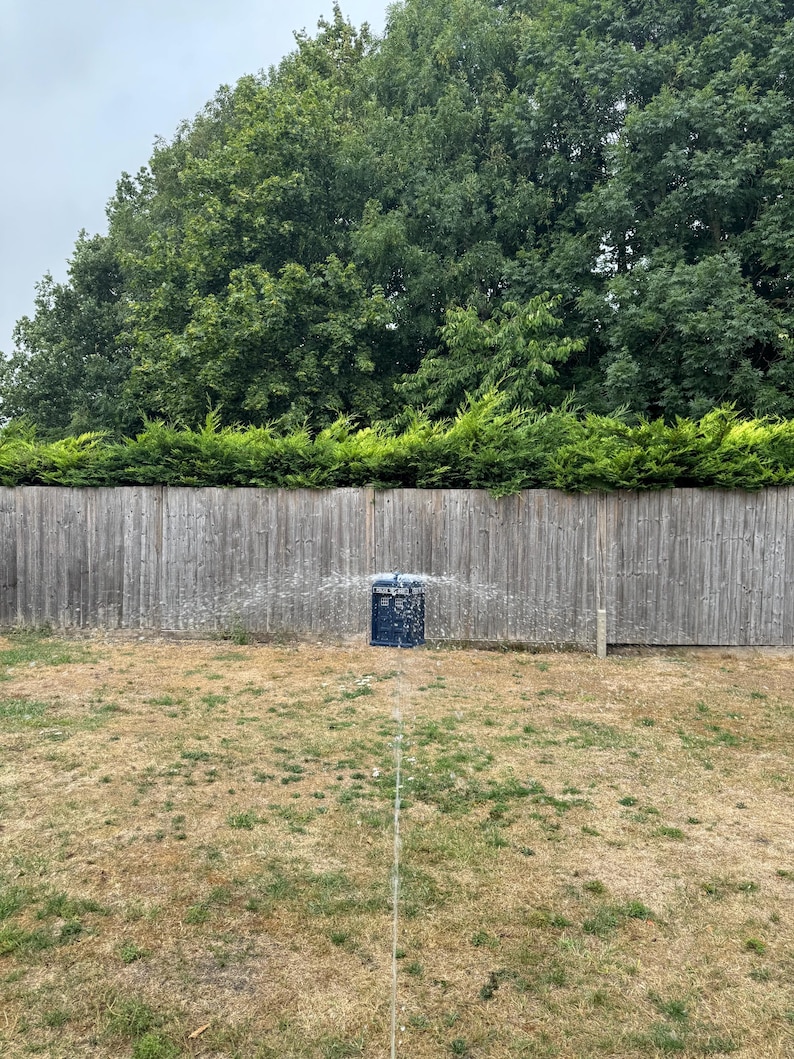 May include: A lawn sprinkler in a yard, spraying water. The sprinkler is black and blue, positioned in front of a wooden fence and green trees. The grass is dry and brown.