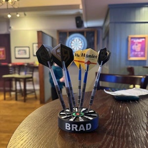 May include: A set of darts in a black and green dart holder with the name "BRAD" in white lettering. The darts have silver shafts and various flight designs, including one with the text "Ha H Hamlet". The darts are on a wooden table.