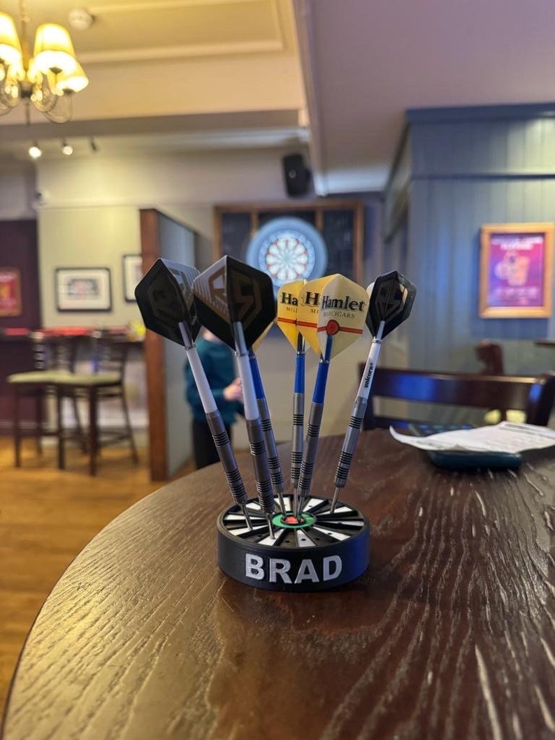 May include: A set of darts in a black and green dart holder with the name "BRAD" in white lettering. The darts have silver shafts and various flight designs, including one with the text "Ha H Hamlet". The darts are on a wooden table.