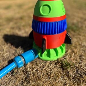 May include: A colorful rocket-shaped lawn sprinkler. The sprinkler features a red, blue, and green design with black fins. A blue hose is attached to the base. The sprinkler is designed to water lawns and gardens.