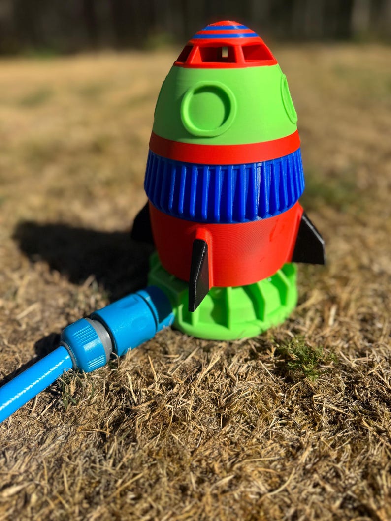 May include: A colorful rocket-shaped lawn sprinkler. The sprinkler features a red, blue, and green design with black fins. A blue hose is attached to the base. The sprinkler is designed to water lawns and gardens.