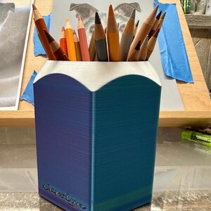 May include: A blue and purple pencil holder with a white top and the name "Carolyn" inscribed on the side. The holder is filled with coloured pencils.