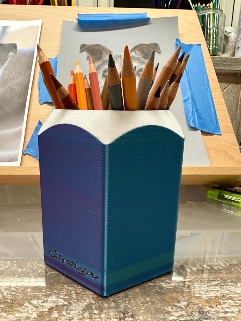 May include: A blue and purple pencil holder with a white top and the name "Carolyn" inscribed on the side. The holder is filled with coloured pencils.