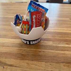 Op de afbeelding: Een witte, voetbalvormige kom gevuld met snoep en lolly's. De kom staat op een zwarte basis met de tekst "DAD'S SNACKS". Het snoepgoed omvat Haribo en Drumstick lolly's. De kom staat op een houten oppervlak.