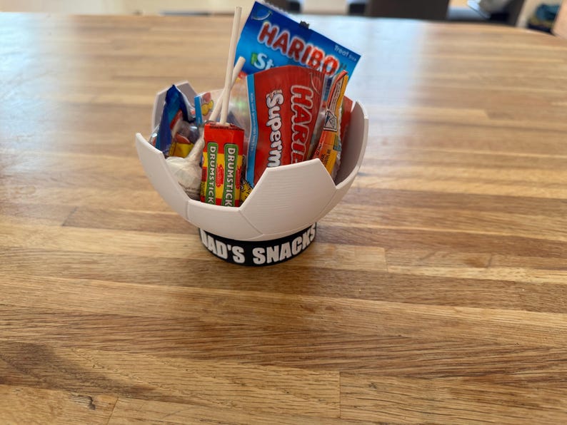 Op de afbeelding: Een witte, voetbalvormige kom gevuld met snoep en lolly's. De kom staat op een zwarte basis met de tekst "DAD'S SNACKS". Het snoepgoed omvat Haribo en Drumstick lolly's. De kom staat op een houten oppervlak.