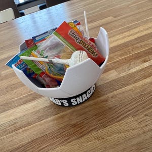 Op de afbeelding: Een witte, voetbalvormige snackkom gevuld met diverse verpakte snoepjes en lollies. De kom staat op een zwarte basis met de tekst "Dad's Snacks" in het wit. Op een houten oppervlak.