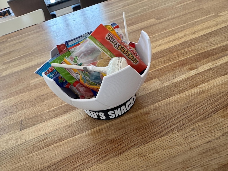 Op de afbeelding: Een witte, voetbalvormige snackkom gevuld met diverse verpakte snoepjes en lollies. De kom staat op een zwarte basis met de tekst "Dad's Snacks" in het wit. Op een houten oppervlak.