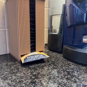 May include: A brown, vertical coffee pod holder with a ribbed design. The holder dispenses individual coffee packets. A blue and white packet of "CAFFE BORBONE" is visible. A blue coffee machine is in the background.