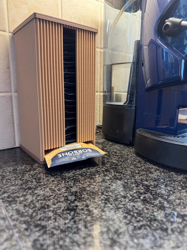 May include: A brown, vertical coffee pod holder with a ribbed design. The holder dispenses individual coffee packets. A blue and white packet of "CAFFE BORBONE" is visible. A blue coffee machine is in the background.