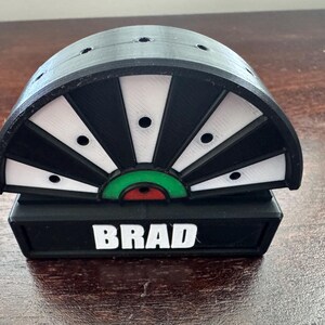 May include: A black and white dartboard with a green and red bullseye. The dartboard is mounted on a black base with the name "BRAD" printed in white.
