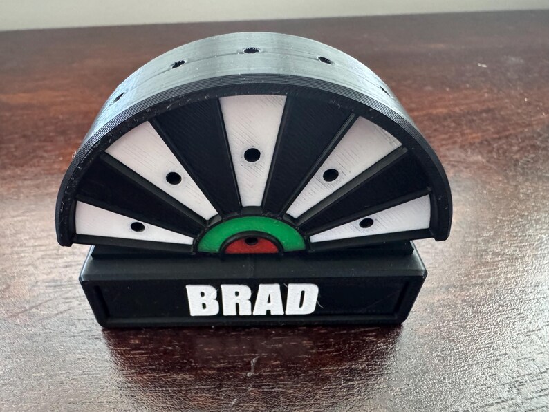 May include: A black and white dartboard with a green and red bullseye. The dartboard is mounted on a black base with the name "BRAD" printed in white.