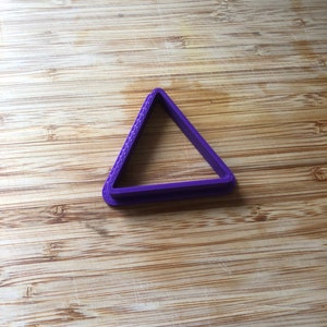 Puede incluir: Cortador de galletas de pl&aacute;stico triangular de color morado sobre una superficie de madera.