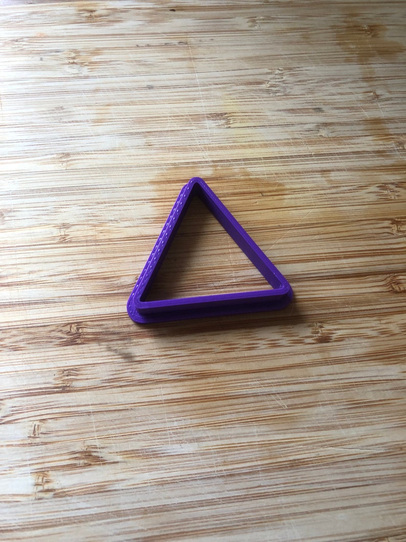Puede incluir: Cortador de galletas de pl&aacute;stico triangular de color morado sobre una superficie de madera.