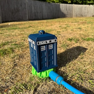 May include: A dark blue Police Box water feature with white text and window details. The box is connected to a green base and a blue hose, set on dry grass. The words "POLICE PUBLIC CALL BOX" are visible on the sign.
