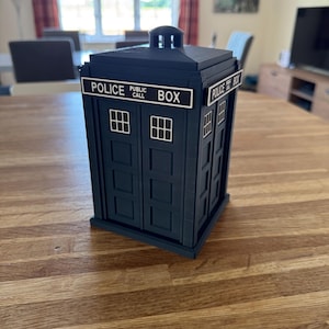 May include: A dark blue Police Box replica with white lettering that reads "POLICE PUBLIC CALL BOX". The box has small, white, rectangular windows and a small, square top. The box is sitting on a wooden table.
