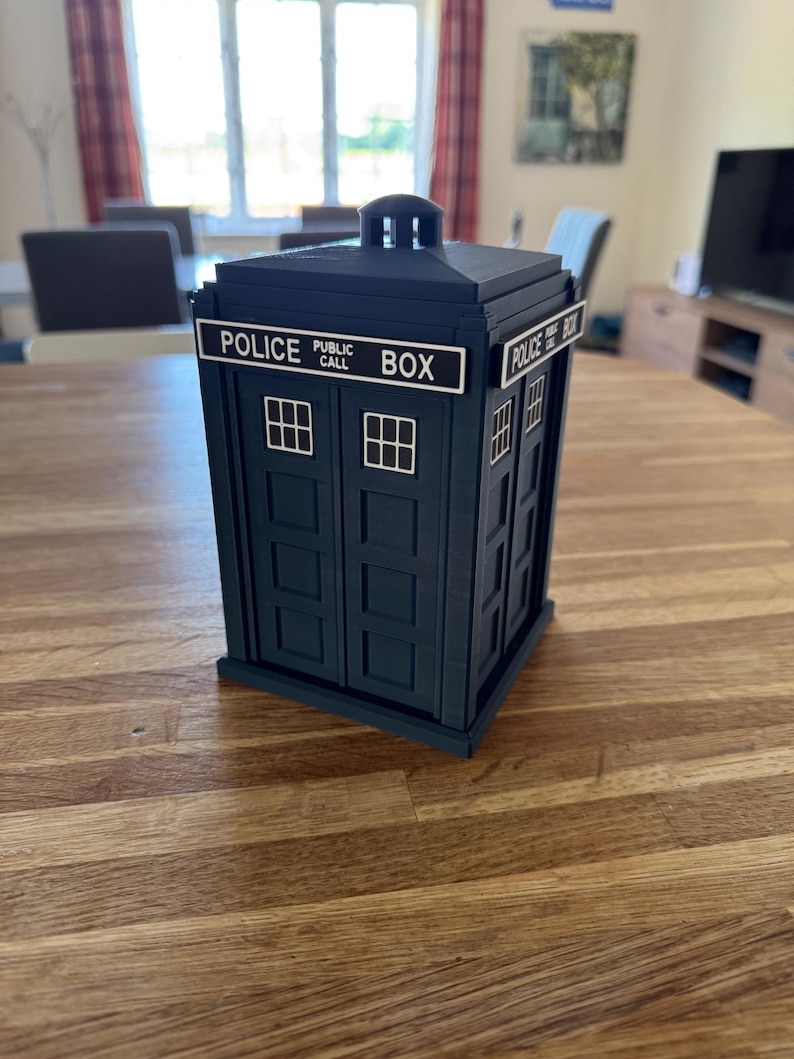 May include: A dark blue Police Box replica with white lettering that reads "POLICE PUBLIC CALL BOX". The box has small, white, rectangular windows and a small, square top. The box is sitting on a wooden table.