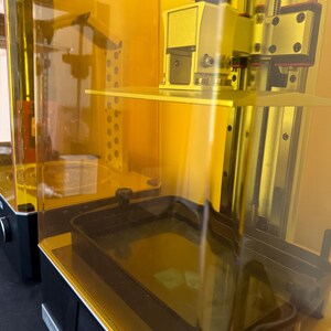 May include: Two yellow resin 3D printers with transparent orange enclosures. The printers have black bases and metallic components. One printer has a black knob and a rectangular build plate. The other printer has a rectangular build plate and a black tray.