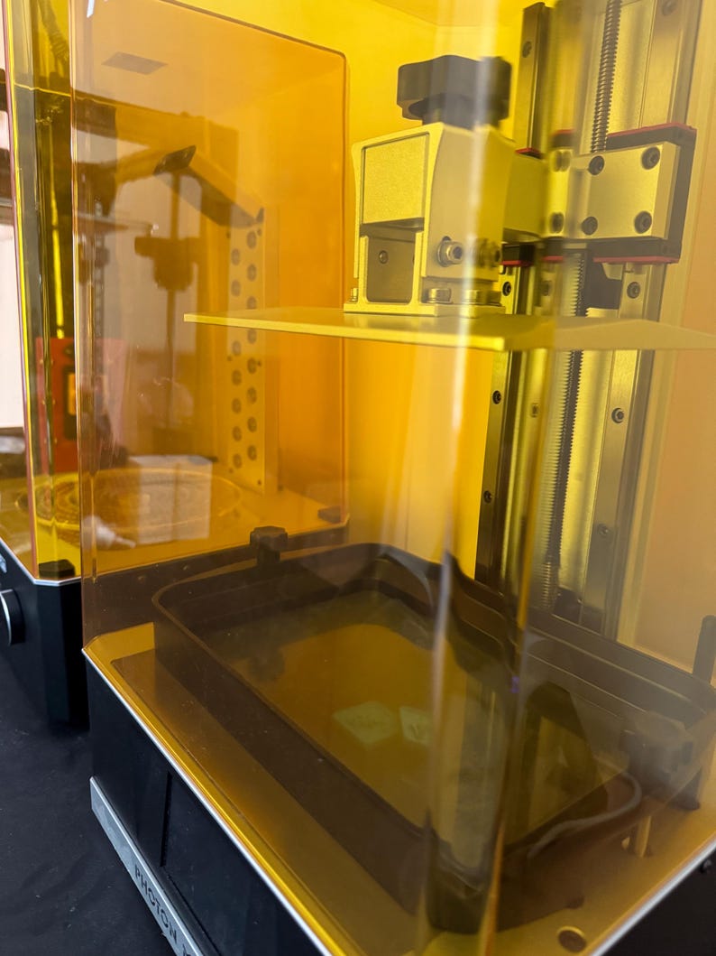 May include: Two yellow resin 3D printers with transparent orange enclosures. The printers have black bases and metallic components. One printer has a black knob and a rectangular build plate. The other printer has a rectangular build plate and a black tray.