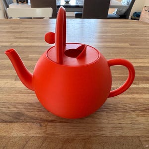 May include: A vibrant red teapot with a rounded body, a curved spout, and a handle. The lid features a unique design with a vertical handle and a small knob. This teapot is designed for brewing tea.