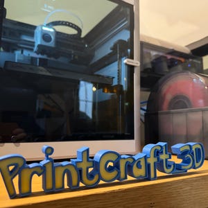 May include: A 3D-printed sign that reads "PrintCraft 3D" in blue and yellow. The sign is on a wooden surface in front of a 3D printer and a spool of red filament.