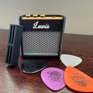 May include: A black and orange miniature guitar amplifier with the word "Lewis" printed on the front. The amplifier is sitting on a wooden surface with four guitar picks next to it.