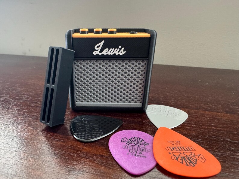 May include: A black and orange miniature guitar amplifier with the word "Lewis" printed on the front. The amplifier is sitting on a wooden surface with four guitar picks next to it.