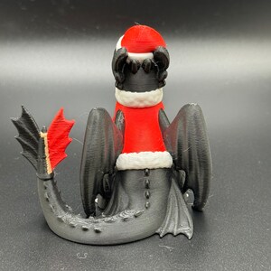 May include: A black 3D printed figurine of a dragon wearing a red and white Santa hat. The dragon is sitting on its hind legs with its tail curled around its body.