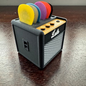 May include: A black miniature guitar amplifier with a speaker grill and a row of knobs. The amplifier has a label that says "Jele" and is holding a set of colorful guitar picks.