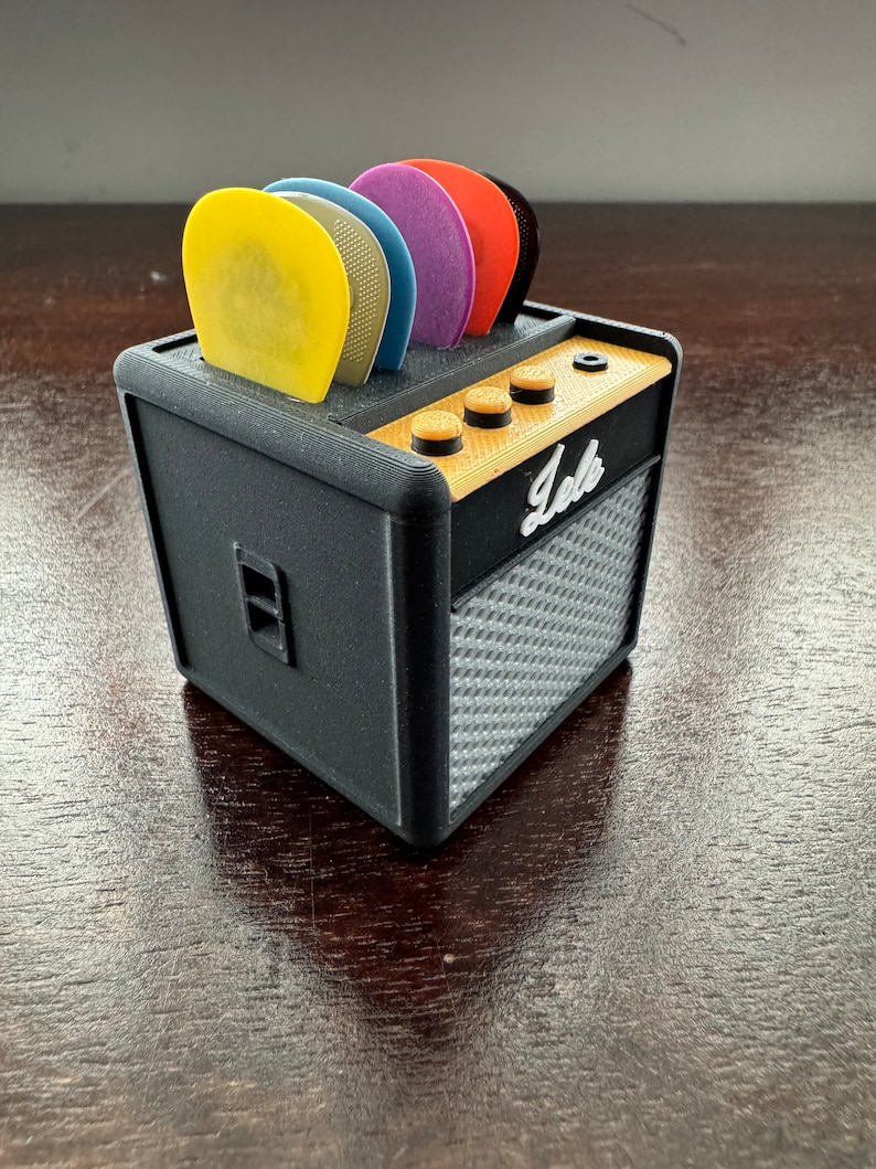 May include: A black miniature guitar amplifier with a speaker grill and a row of knobs. The amplifier has a label that says "Jele" and is holding a set of colorful guitar picks.
