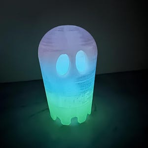 Op de afbeelding: Een gloeiend spookvormig nachtlampje. Het licht gaat over van een groene basis naar blauw en paars aan de bovenkant. De geest heeft twee ovale ooguitsparingen. Het product is ontworpen om in het donker te gloeien.