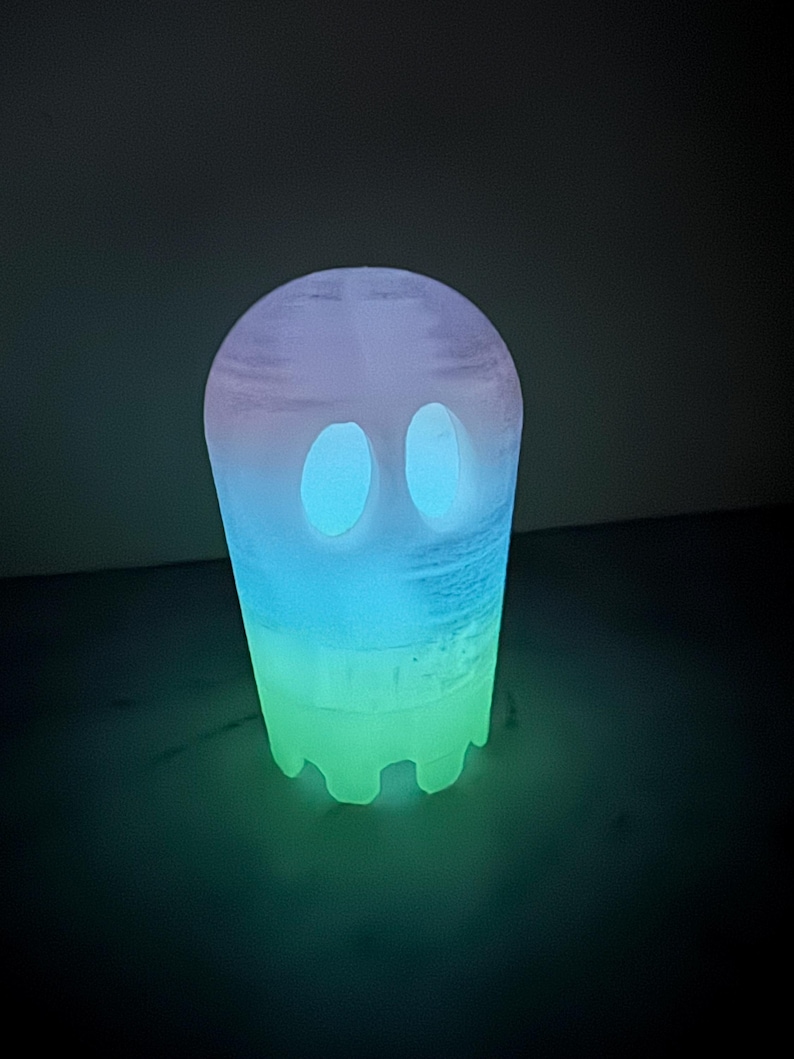 Op de afbeelding: Een gloeiend spookvormig nachtlampje. Het licht gaat over van een groene basis naar blauw en paars aan de bovenkant. De geest heeft twee ovale ooguitsparingen. Het product is ontworpen om in het donker te gloeien.