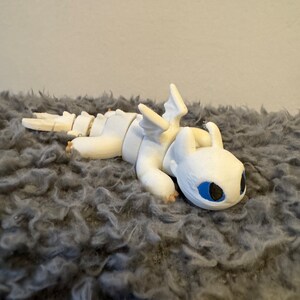 May include: White articulated dragon figurine with blue eyes, lying on a gray fluffy surface.