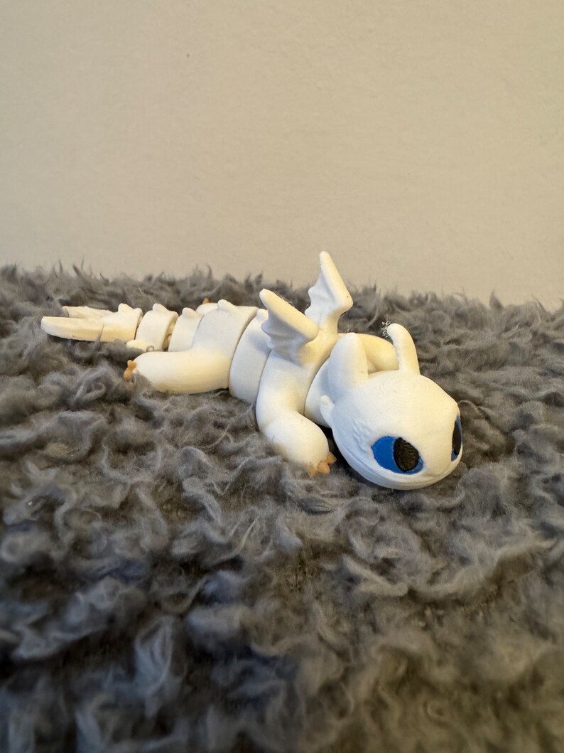 May include: White articulated dragon figurine with blue eyes, lying on a gray fluffy surface.