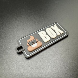 May include: A black and white rectangular tag with a brown poop emoji wearing sunglasses and the word "BOX" in white letters.