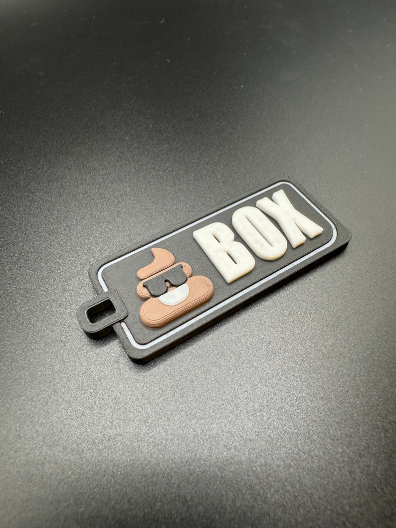 May include: A black and white rectangular tag with a brown poop emoji wearing sunglasses and the word "BOX" in white letters.