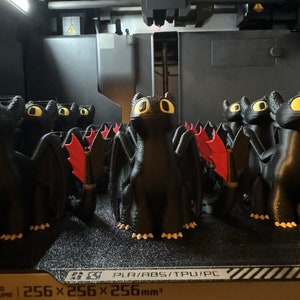 May include: A 3D printer printing a row of black dragon figurines with red wings. The printer bed is labeled "256 x 256 x 256 mm3" and "PLA/ABS/TPU/PC".