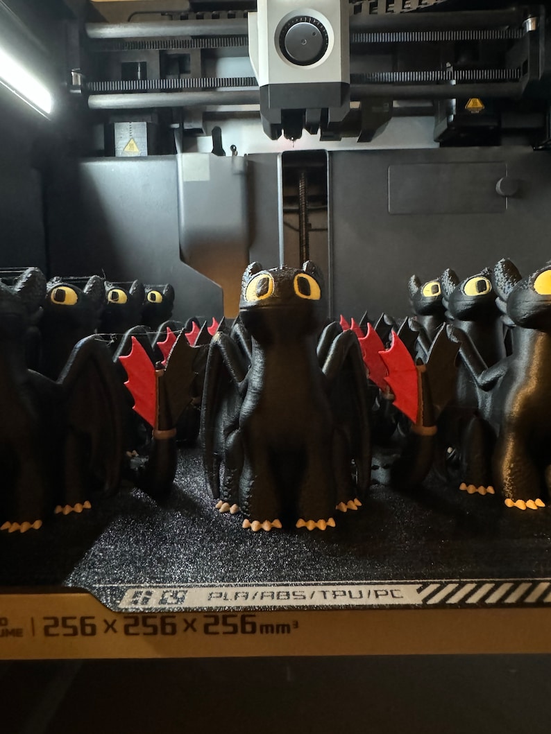 May include: A 3D printer printing a row of black dragon figurines with red wings. The printer bed is labeled "256 x 256 x 256 mm3" and "PLA/ABS/TPU/PC".