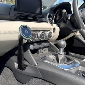 May include: Interior view of a car's dashboard, featuring a central display screen, air conditioning controls, and a gear shift. A circular device with a blue and green design is mounted on a black stand. The car's interior is primarily black and cream.