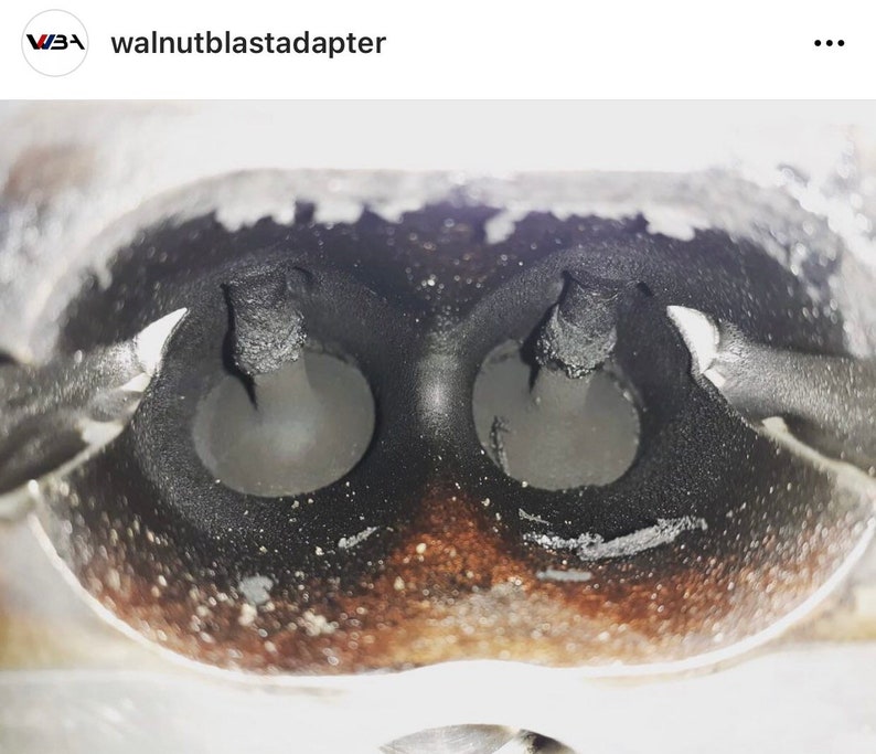 May include: Close-up of a dirty engine intake manifold with two round ports. The ports are covered in black soot and debris. The manifold is made of metal and has a rough, textured surface.