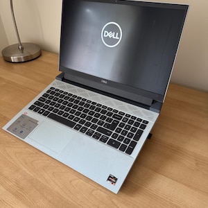 May include: A Dell laptop with a black screen displaying the Dell logo. The laptop has a black keyboard and a light gray palm rest. The laptop is open on a wooden desk.