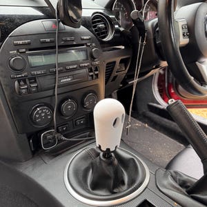 Op de afbeelding: Een witte, spookvormige versnellingspookknop in een auto. De knop heeft twee oogvormige uitsparingen en is gemonteerd op een zwarte versnellingspook. Het interieur van de auto heeft een zwart dashboard, stereo-installatie en ventilatieopeningen.