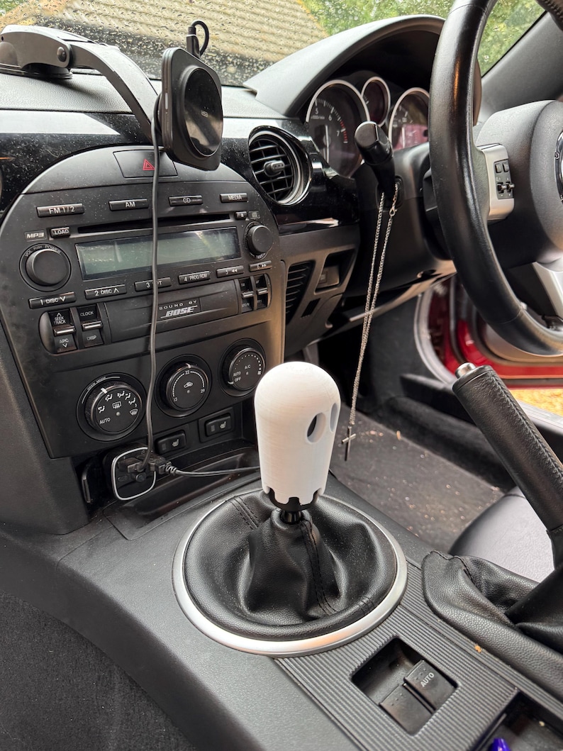 Op de afbeelding: Een witte, spookvormige versnellingspookknop in een auto. De knop heeft twee oogvormige uitsparingen en is gemonteerd op een zwarte versnellingspook. Het interieur van de auto heeft een zwart dashboard, stereo-installatie en ventilatieopeningen.