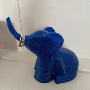 Op de afbeelding: Een blauwe glitter olifant ringhouder. De olifant heeft een opgetrokken slurf en staat op een wit oppervlak.