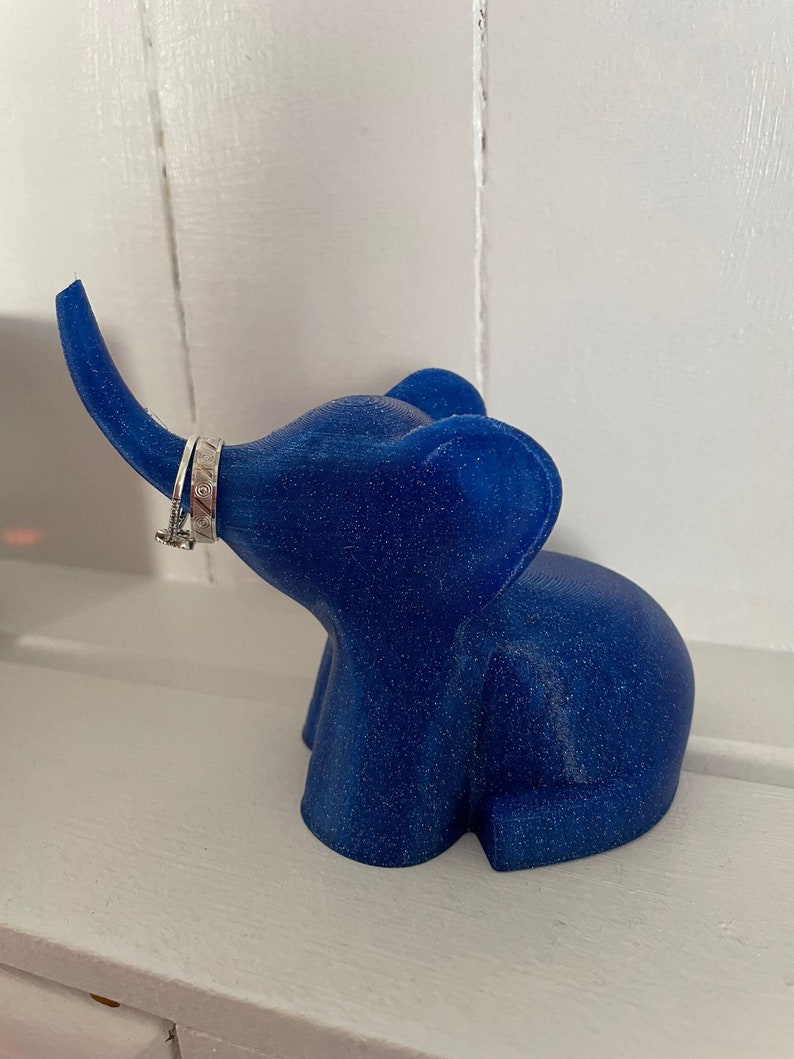Op de afbeelding: Een blauwe glitter olifant ringhouder. De olifant heeft een opgetrokken slurf en staat op een wit oppervlak.